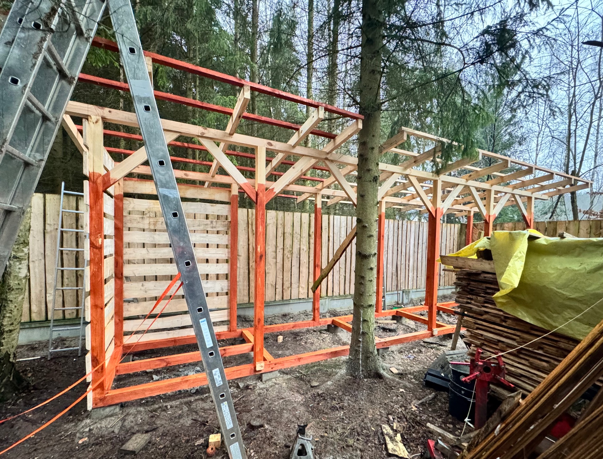 Konstrukcja drewniana w trakcie budowy, pomalowane na czerwono słupy i belki, widoczne deski i drabina, drzewa w tle. Budowa w toku, teren budowy.