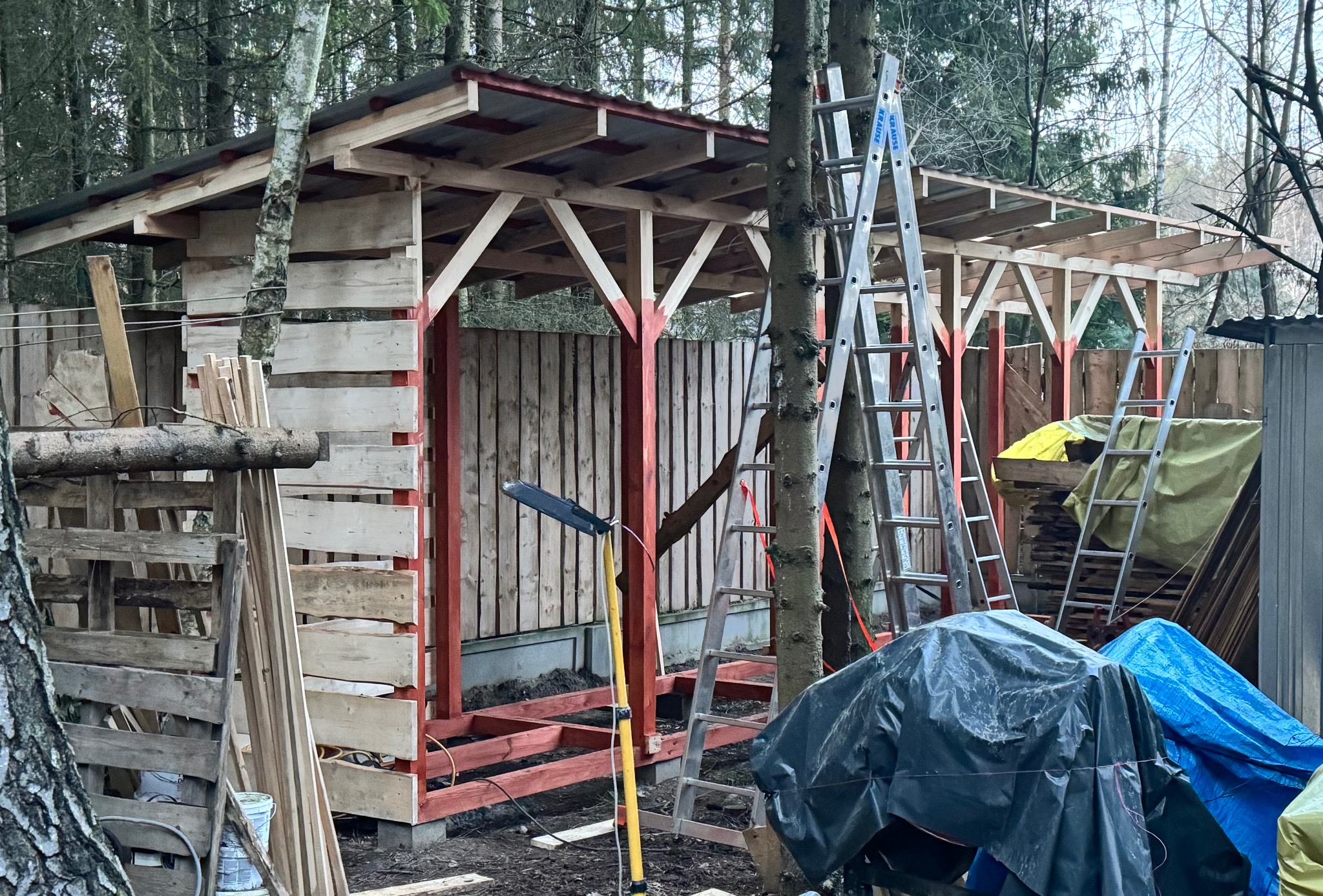 Konstrukcja z drewna w trakcie budowy, częściowo obita deskami, z czerwonymi elementami. W tle las, na pierwszym planie drabiny i materiały budowlane.