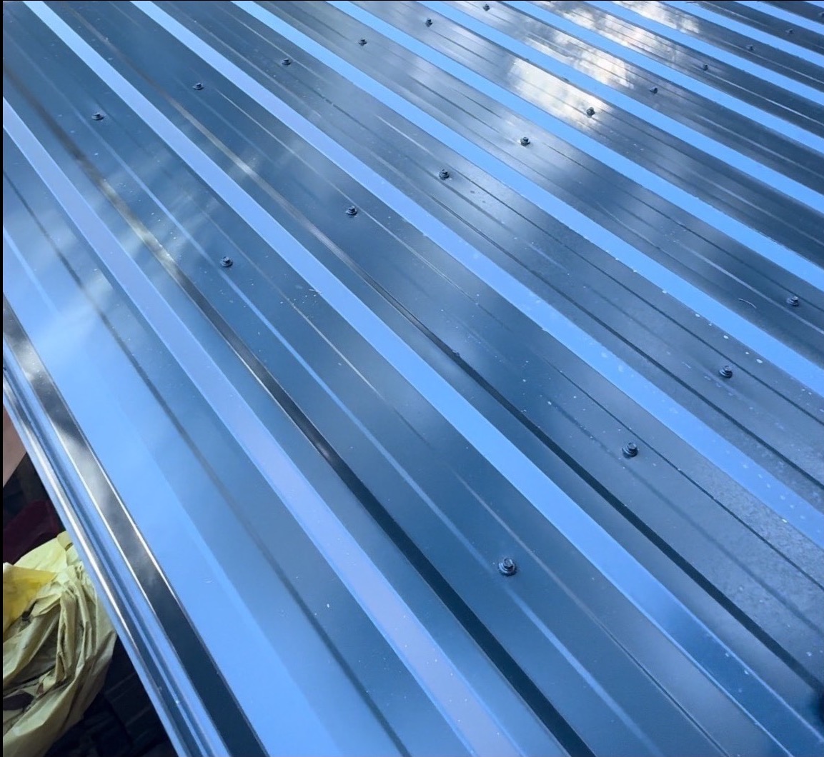 Szare, metalowe pokrycie dachowe z widocznymi śrubami mocującymi, ukazane pod kątem. Widoczna faktura blachy i odbicia światła.