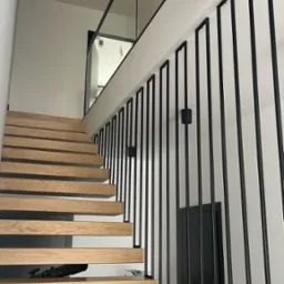 Nowoczesne schody drewniane z czarną metalową balustradą o minimalistycznym designie i szklanym panelem na górze, oświetlone punktowymi lampami.