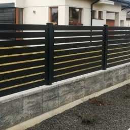 Energo Usługi Spawalnicze GRZEGORZ GODULA - Nowoczesne ogrodzenie metalowe z poziomymi, czarnymi panelami i złotymi akcentami, osadzone na niskim murku z szarych bloczków. Tło: domy jednorodzinne i szara nawierzchnia.