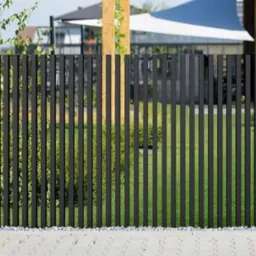 Nowoczesne ogrodzenie z czarnych, pionowych sztachet metalowych, osadzone między betonowymi słupkami. W tle zieleń trawnika i elementy architektury.