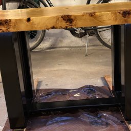 Energo Usługi Spawalnicze GRZEGORZ GODULA - Stolik z blatem z jasnego drewna z wyraźnymi słojami i czarną, metalową podstawą. W tle widoczne rower i ściana. Całość na folii ochronnej.