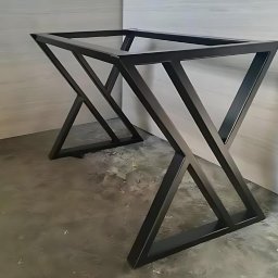 Energo Usługi Spawalnicze GRZEGORZ GODULA - Metalowa podstawa stołu o geometrycznym kształcie X, pomalowana na czarno, ustawiona na szarej posadzce przy białej ścianie, nowoczesny design.