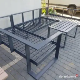 Metalowy zestaw mebli ogrodowych: sofa i stolik, pomalowane na szaro, z ażurowymi siedziskami i oparciami. Ustawione na kostce brukowej, w tle biała ściana.