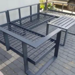 Metalowy stelaż sofy i stolika na tarasie, konstrukcja spawana z profili, pomalowana na szaro, widoczne detale łączeń i regularny układ szczebli.