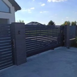Energo Usługi Spawalnicze GRZEGORZ GODULA - Nowoczesna, przesuwna brama wjazdowa z poziomymi panelami w kolorze grafitowym, zamontowana przy posesji z jasnym budynkiem w tle. Widoczne słupki i elementy automatyki.