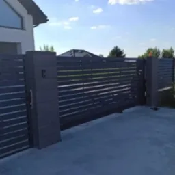 Nowoczesna, przesuwna brama wjazdowa z poziomymi panelami w kolorze grafitowym, zamontowana przy posesji z jasnym budynkiem w tle. Widoczne słupki i elementy automatyki.