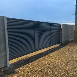 Energo Usługi Spawalnicze GRZEGORZ GODULA - Nowoczesna, przesuwna brama wjazdowa z lamelami w kolorze antracytu, zamontowana między betonowymi słupkami ogrodzeniowymi. Widok z zewnątrz, w tle drzewo.