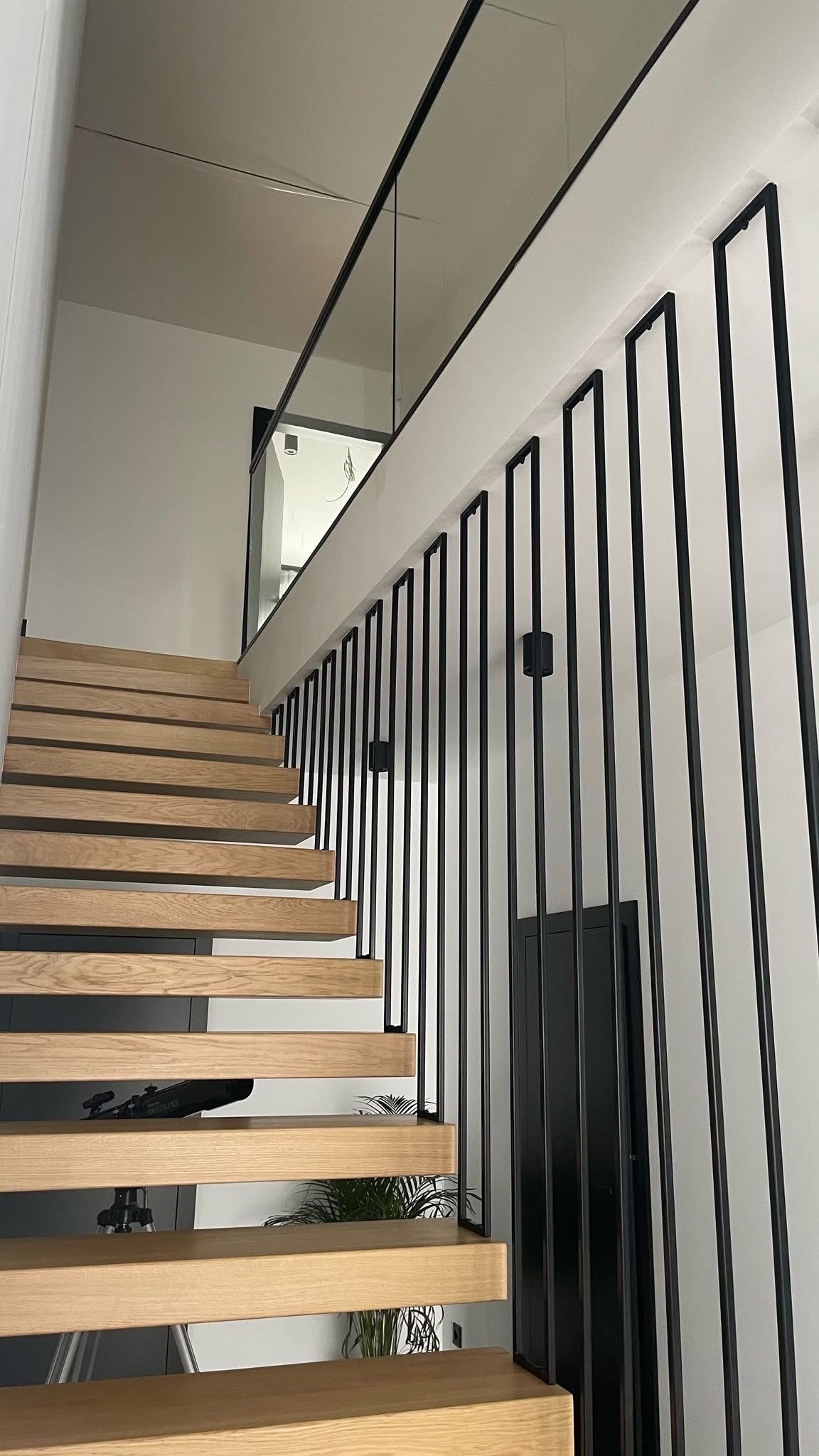 Nowoczesne schody drewniane z czarną metalową balustradą o minimalistycznym designie i szklanym panelem na górze, oświetlone punktowymi lampami.