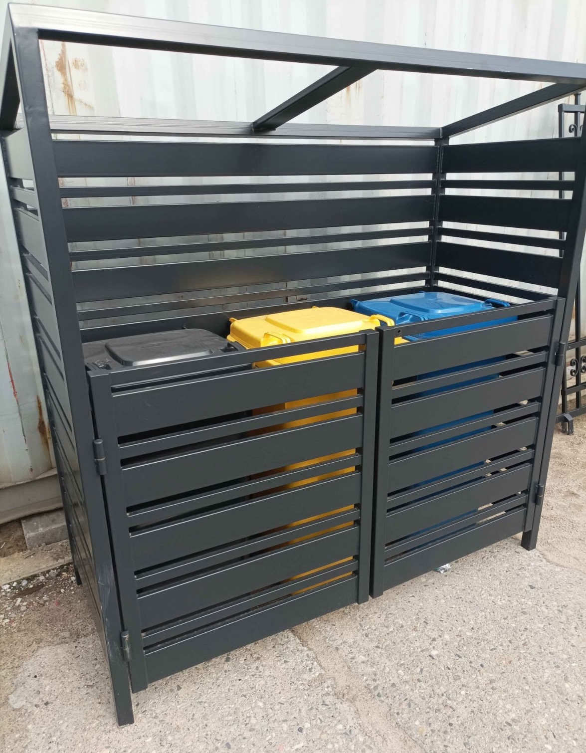 Metalowa zabudowa śmietnikowa w kolorze grafitowym z poziomymi szczeblami, skrywająca pojemniki na odpady w kolorach: czarnym, żółtym i niebieskim.