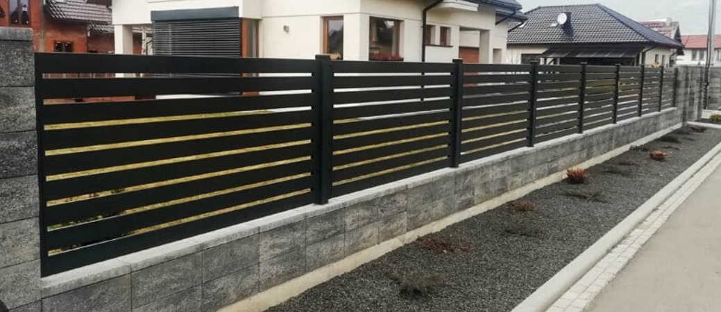 Nowoczesne ogrodzenie metalowe z poziomymi, czarnymi panelami i złotymi akcentami, osadzone na niskim murku z szarych bloczków. Tło: domy jednorodzinne i szara nawierzchnia.