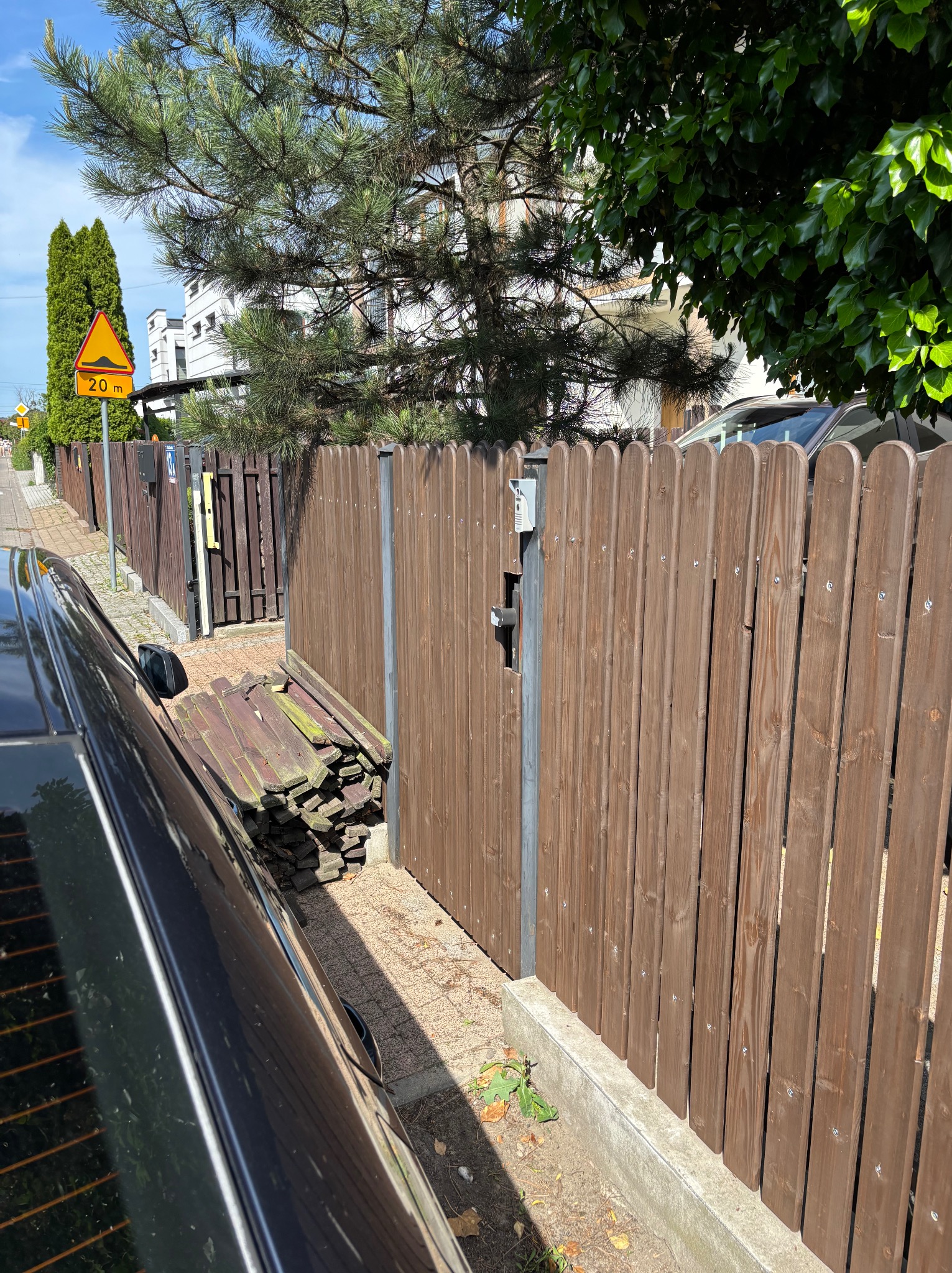 Drewniane ogrodzenie panelowe z furtką i stertą starych desek obok, widok zza zaparkowanego samochodu na tle zabudowy jednorodzinnej i znaku drogowego.