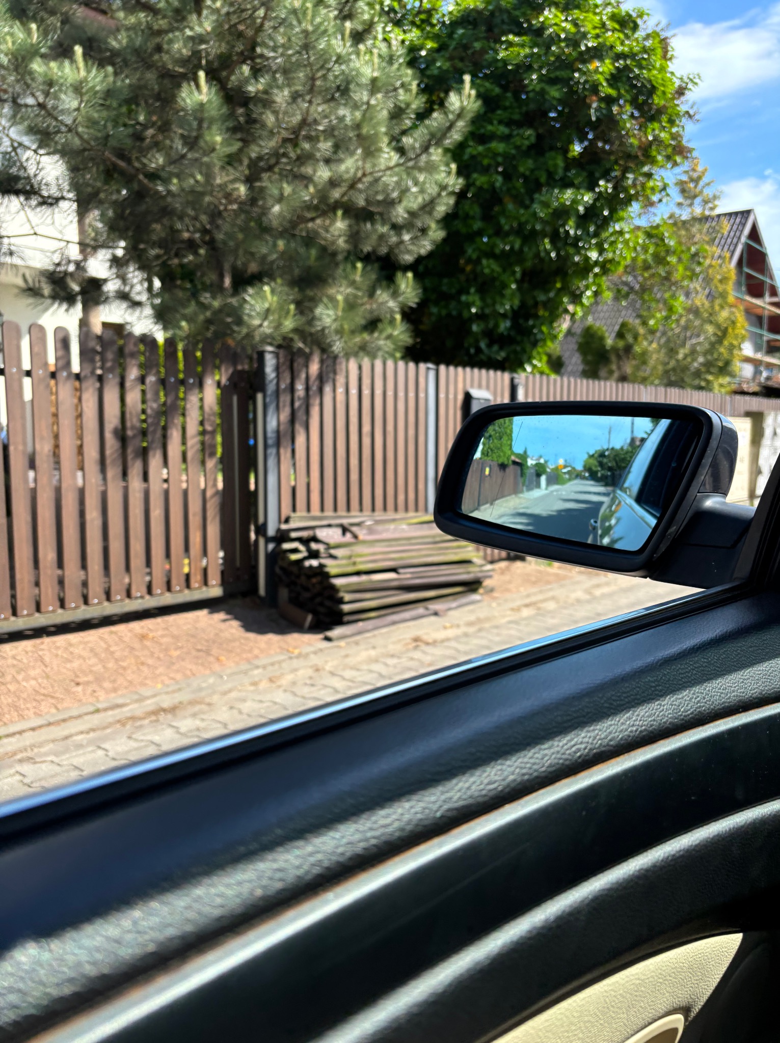 Widok z wnętrza samochodu na brązowe ogrodzenie drewniane i stos desek obok, odbicie ulicy w lusterku bocznym.