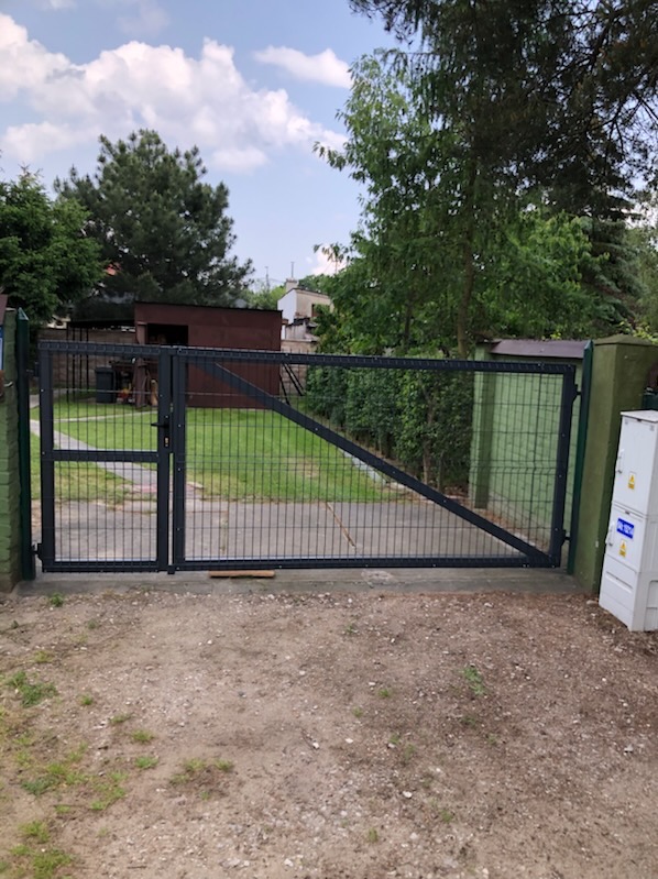 Nowoczesna, grafitowa brama panelowa z furtką, z ukośnym wzmocnieniem, zamontowana na betonowej podmurówce. W tle zieleń drzew i zabudowania.