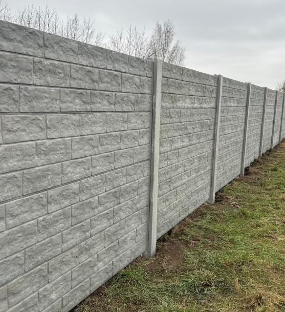 Długi, szary mur z prefabrykatów betonowych, imitujących cegłę, z widocznymi słupkami, ciągnący się wzdłuż trawnika. Tło stanowią drzewa i pochmurne niebo.