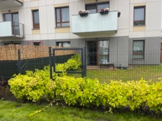 Nowoczesne ogrodzenie panelowe z podkładem z trzciny, obsadzone dekoracyjną roślinnością, na tle budynku wielorodzinnego z balkonami.