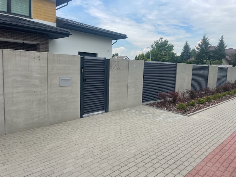 Nowoczesne ogrodzenie z szarych, poziomych paneli i betonowych słupków, brama wejściowa z numerem domu '81', skrzynka pocztowa, kostka brukowa.