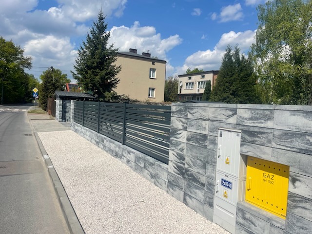 Nowoczesne ogrodzenie z poziomych paneli metalowych, osadzone na murze z szarych bloczków imitujących kamień, z żółtą skrzynką gazową w tle, widok z chodnika.