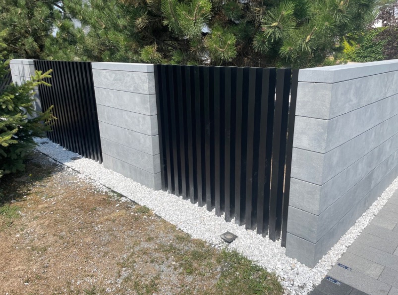 Nowoczesne ogrodzenie z szarych betonowych bloczków i czarnych, pionowych paneli, oddzielone białym żwirem. Widok na fragment posesji z zielenią.