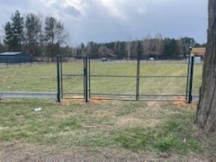 Nowoczesne ogrodzenie panelowe z bramą wjazdową na posesji, widok z poziomu gruntu, z fragmentem drzewa w kadrze i budynkiem w tle.