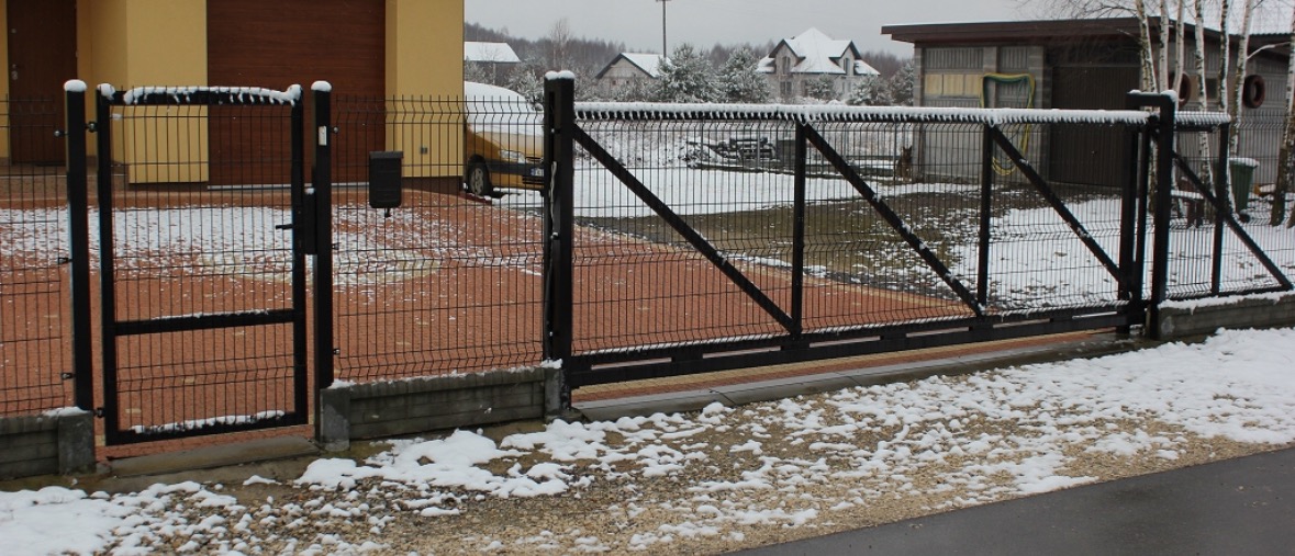 Nowoczesna, czarna brama przesuwna i furtka z paneli ogrodzeniowych na posesji w zimowej scenerii. Elementy konstrukcyjne pokryte śniegiem.