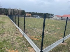 Długie, panelowe ogrodzenie w kolorze grafitowym z podmurówką betonową, otaczające rozległy teren zielony. W tle zabudowania mieszkalne i las.