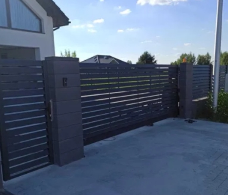 Nowoczesna, przesuwna brama wjazdowa z poziomymi panelami w kolorze grafitowym, zamontowana przy posesji z jasnym budynkiem w tle. Widoczne słupki i elementy automatyki.