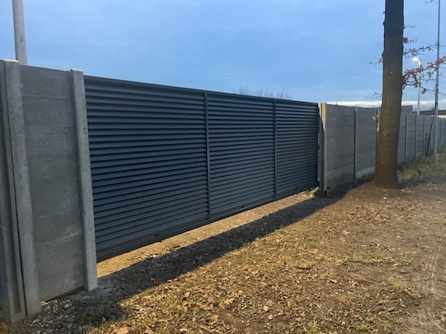 Nowoczesna, przesuwna brama wjazdowa z lamelami w kolorze antracytu, zamontowana między betonowymi słupkami ogrodzeniowymi. Widok z zewnątrz, w tle drzewo.