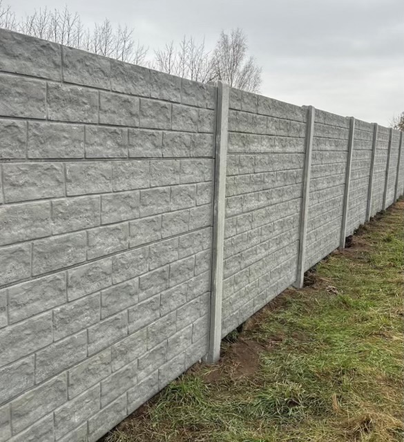 Długi, szary, betonowy płot panelowy z fakturą cegły, widziany z perspektywy ziemi. Słupki pionowe, trawa u podnóża, drzewa w tle pod pochmurnym niebem.
