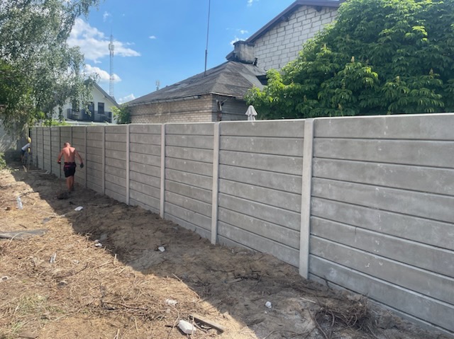 Nowoczesne, betonowe ogrodzenie panelowe z poziomymi żłobieniami, świeżo zamontowane wzdłuż działki. Widoczny monter w tle. Trawa i drzewa w tle.