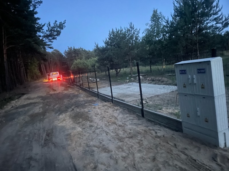 Nowoczesne ogrodzenie panelowe z podmurówką na nierównym terenie leśnym, skrzynka elektryczna obok, samochód dostawczy w tle. Ujęcie z dołu, wieczorne światło.