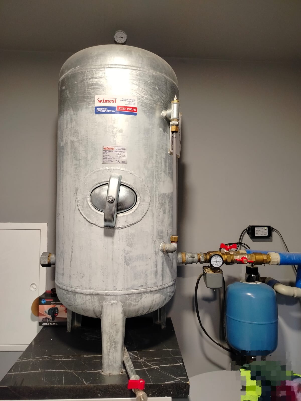 Szary zbiornik hydroforowy Wimest ZCS/150/6 z armaturą i manometrem na ciemnym blacie, widok z przodu. Element instalacji w pomieszczeniu o szarych ścianach.