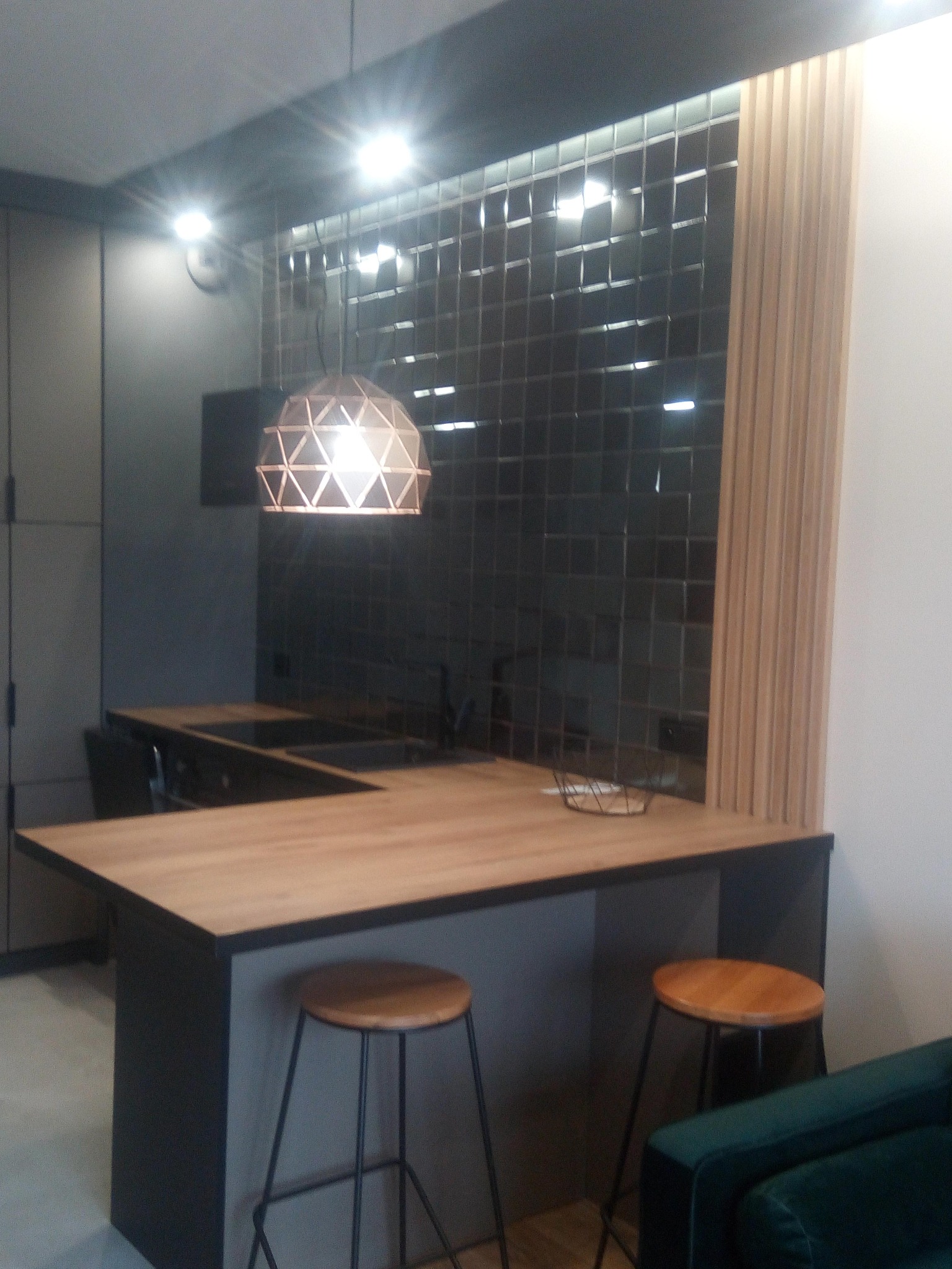 Nowoczesna kuchnia z ciemnymi płytkami i drewnianym blatem. Dwa hokery stoją przy wyspie, nad którą wisi geometryczna lampa. Widoczny fragment zielonej kanapy.