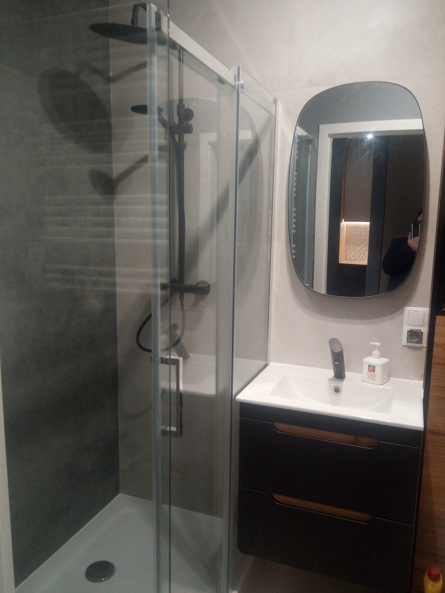 Nowoczesna łazienka z kabiną prysznicową typu walk-in, umywalką z szafką w kolorze ciemnego drewna i owalnym lustrem. Szare płytki na ścianach tworzą stonowane tło.