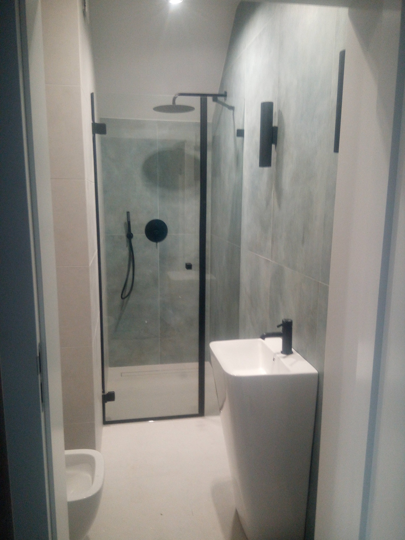 Nowoczesna łazienka z kabiną prysznicową typu walk-in, umywalką wolnostojącą i szarą glazurą. Minimalistyczne czarne akcenty armatury i oświetlenia.