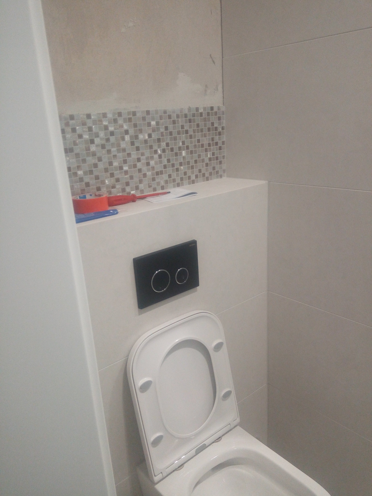 Wykończona toaleta z mozaiką i zabudową geberit. Widoczne narzędzia, co sugeruje prace wykończeniowe. Minimalistyczny design z szarą kolorystyką.