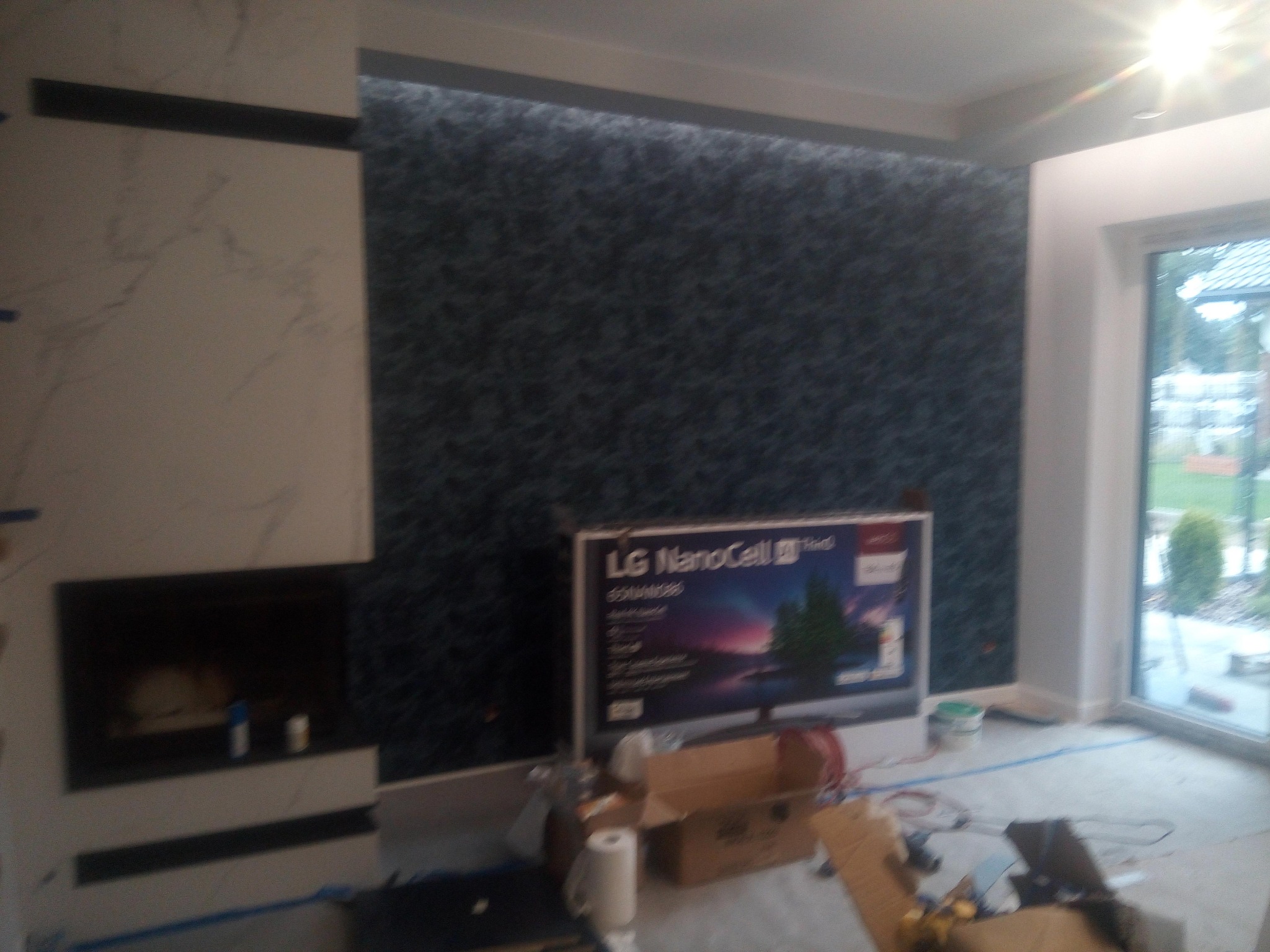 Fragment pokoju z kominkiem w trakcie wykończenia: ciemna tapeta na ścianie, obudowa kominka z jasnego marmuru, widoczne narzędzia i materiały budowlane.