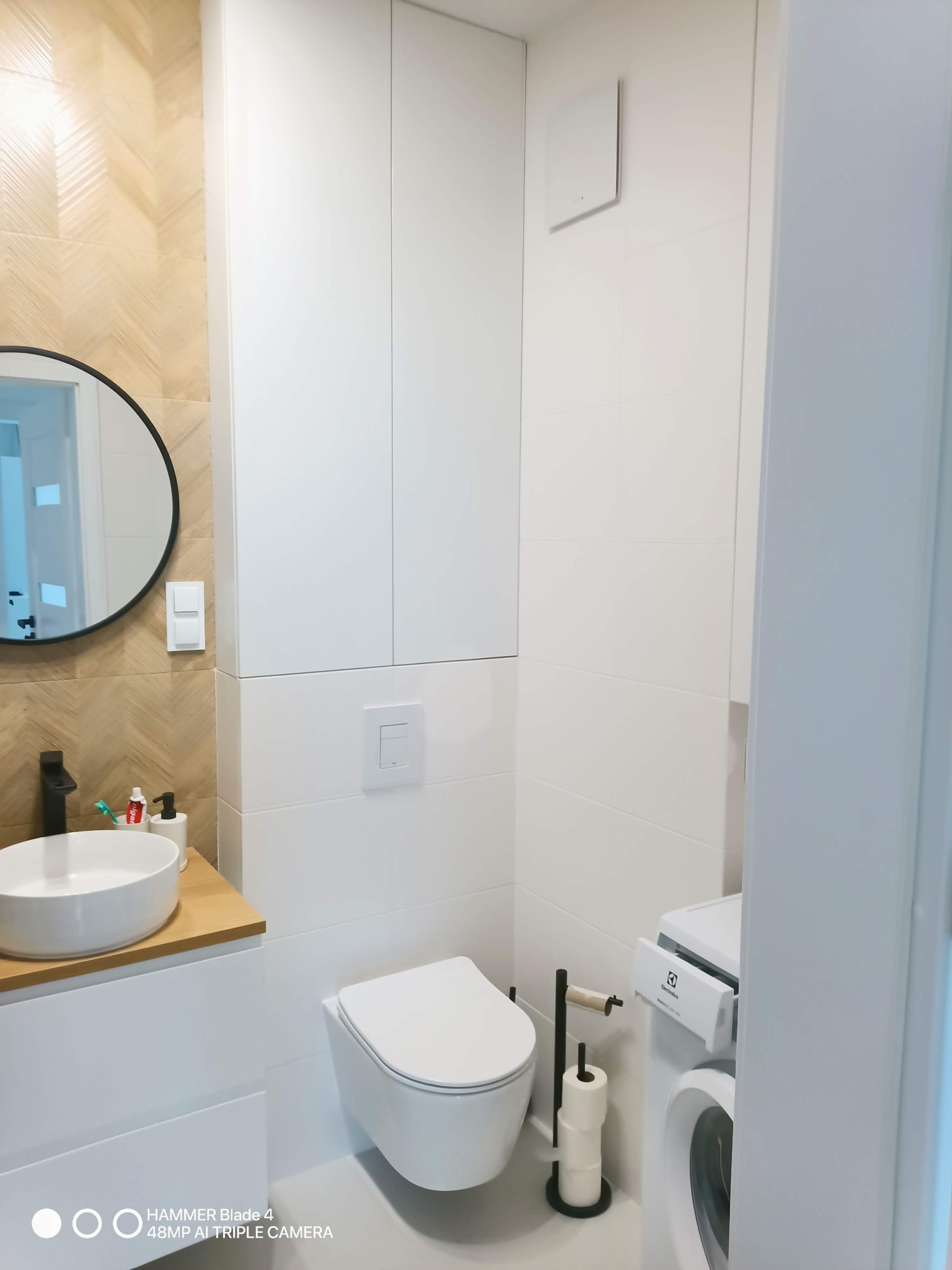 Nowoczesna łazienka w bieli i beżu: okrągłe lustro, umywalka na blacie, podwieszana toaleta i pralka. Ściana wykończona płytkami w jodełkę.