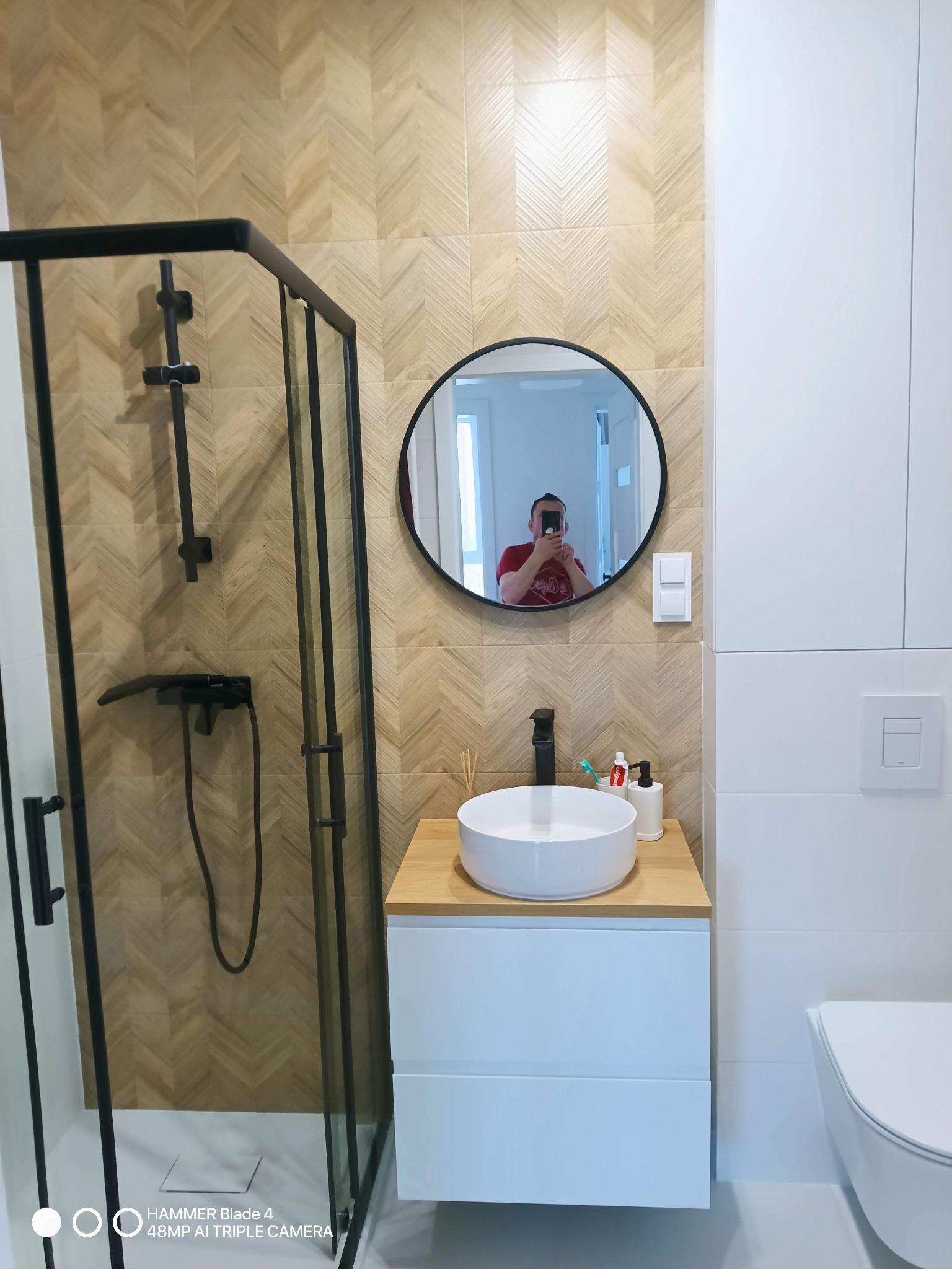 Nowoczesna łazienka z kabiną prysznicową typu walk-in, umywalką nablatową i lustrem w czarnej ramie. Ściany wyłożone płytkami w jodełkę.