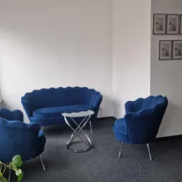 Niebieski, welurowy komplet wypoczynkowy (sofa i dwa fotele) z okrągłym stolikiem na metalowej podstawie w minimalistycznym biurze z obrazkami na ścianie.