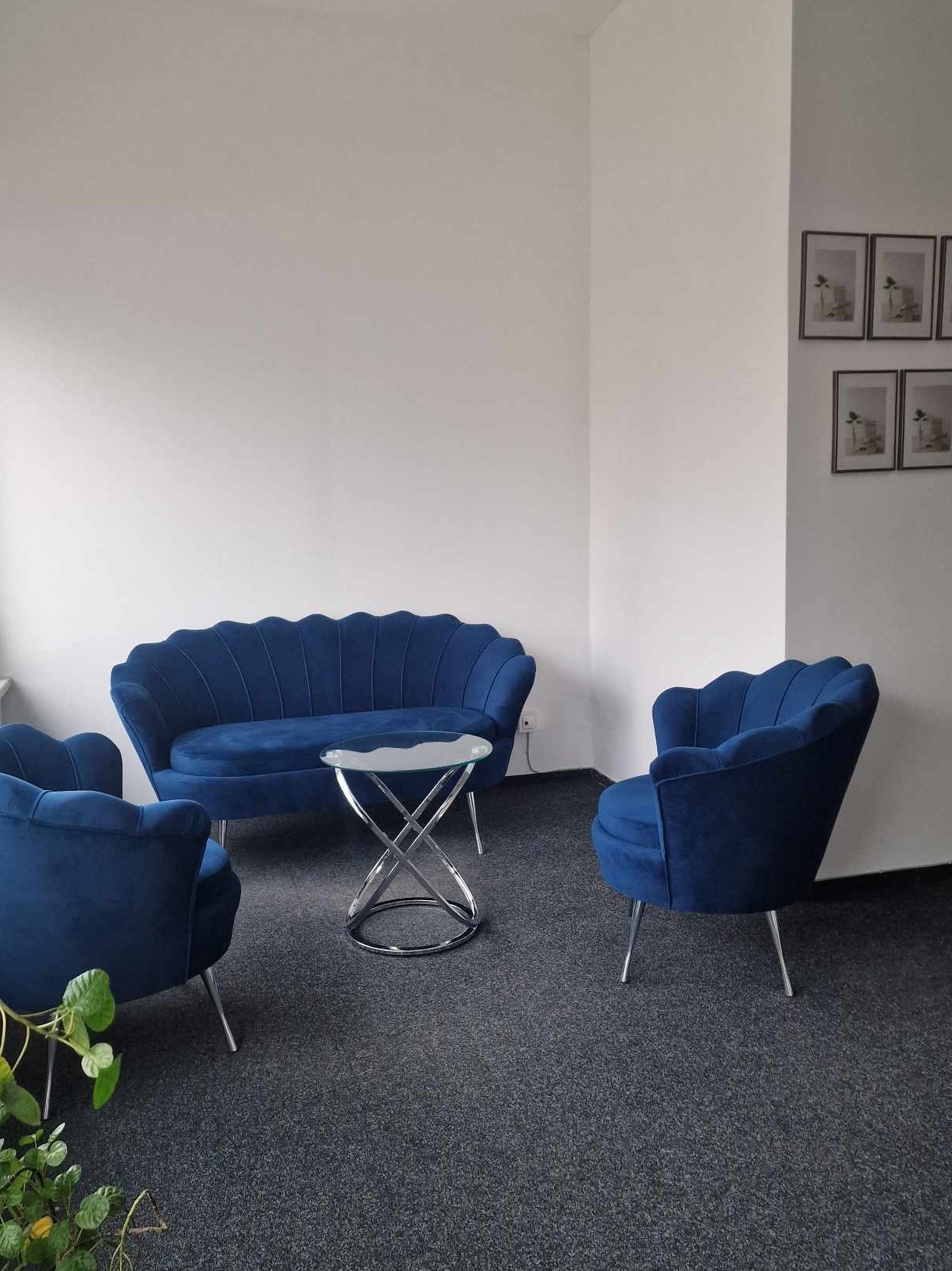 Niebieski, welurowy komplet wypoczynkowy (sofa i dwa fotele) z okrągłym stolikiem na metalowej podstawie w minimalistycznym biurze z obrazkami na ścianie.