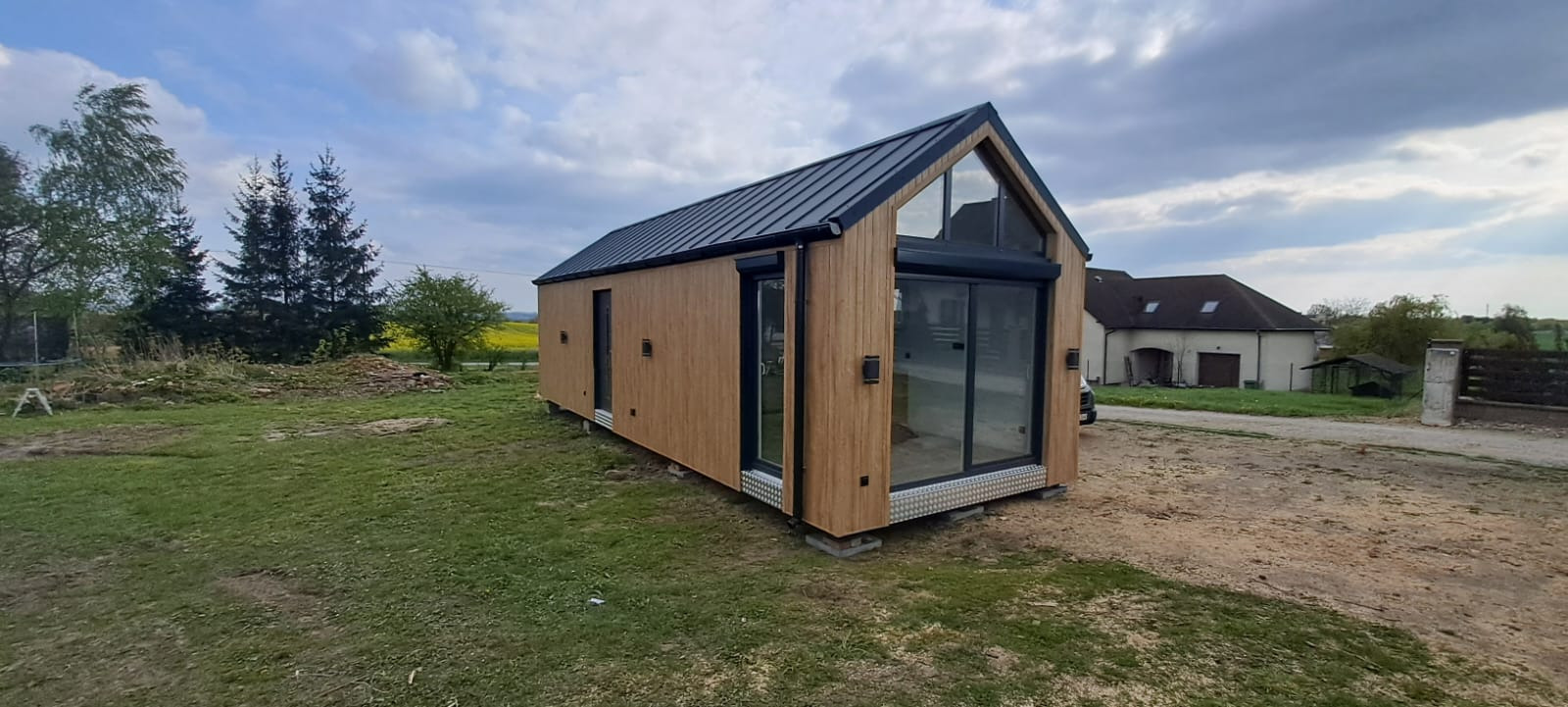 Gotowy dom modułowy z drewnianą elewacją, dużymi oknami i czarnym dachem na zielonej działce, w tle budynek gospodarczy i drzewa.