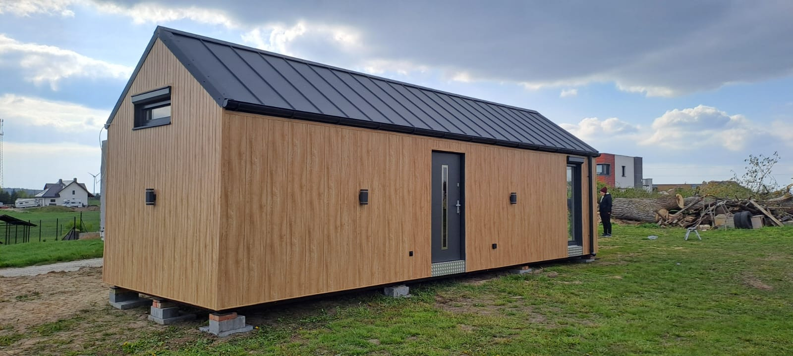 Gotowy, parterowy dom z drewnianą elewacją i czarnym dachem, posadowiony na betonowych bloczkach. W tle osoba i stos pociętych pni drzew.