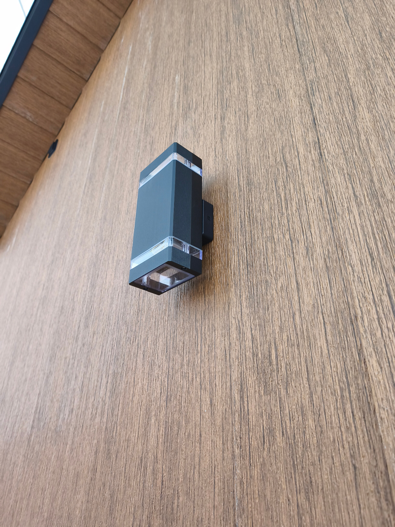 Elegancki, czarny kinkiet z przezroczystymi akcentami zamontowany na drewnianej elewacji domu modułowego. Minimalistyczny design oświetlenia zewnętrznego.