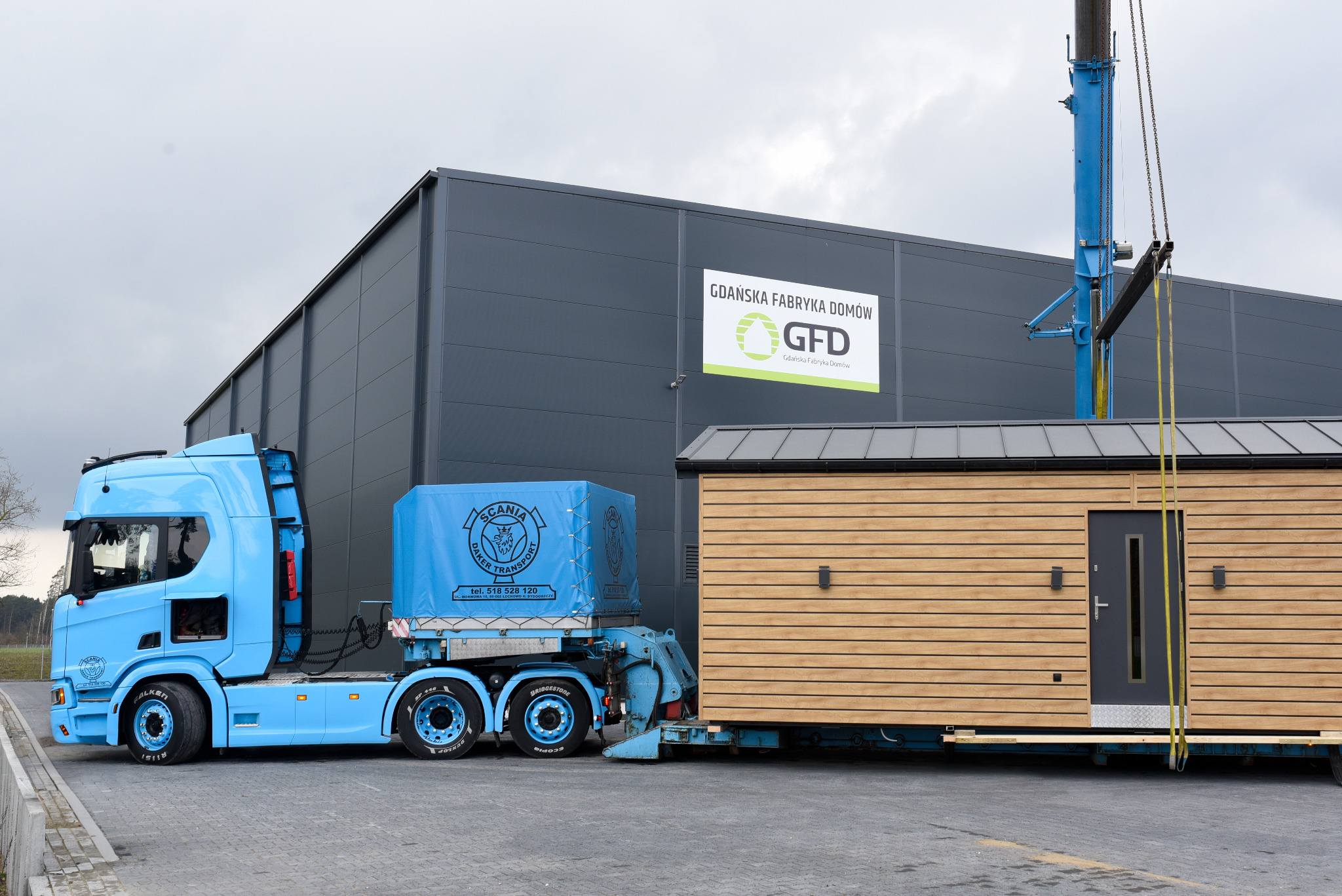 Błękitna ciężarówka Scania z modułem domu na naczepie, na tle szarego budynku fabryki domów GFD. Żuraw podnosi moduł. Widok z boku, pochmurne niebo.