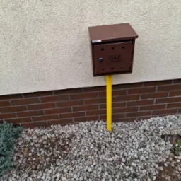 Brązowa skrzynka gazowa z żółtą rurą na tle beżowej ściany i brązowej cegły, otoczona białymi kamieniami i zielonymi roślinami. Widoczne elementy instalacji.