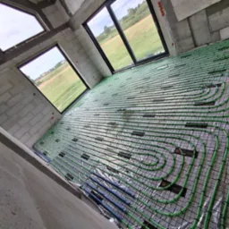 Ogrzewanie podłogowe w surowym wnętrzu w Rąbiu: zielone rurki na czarnej siatce, widok z góry, okna z widokiem na zewnątrz, surowy beton.