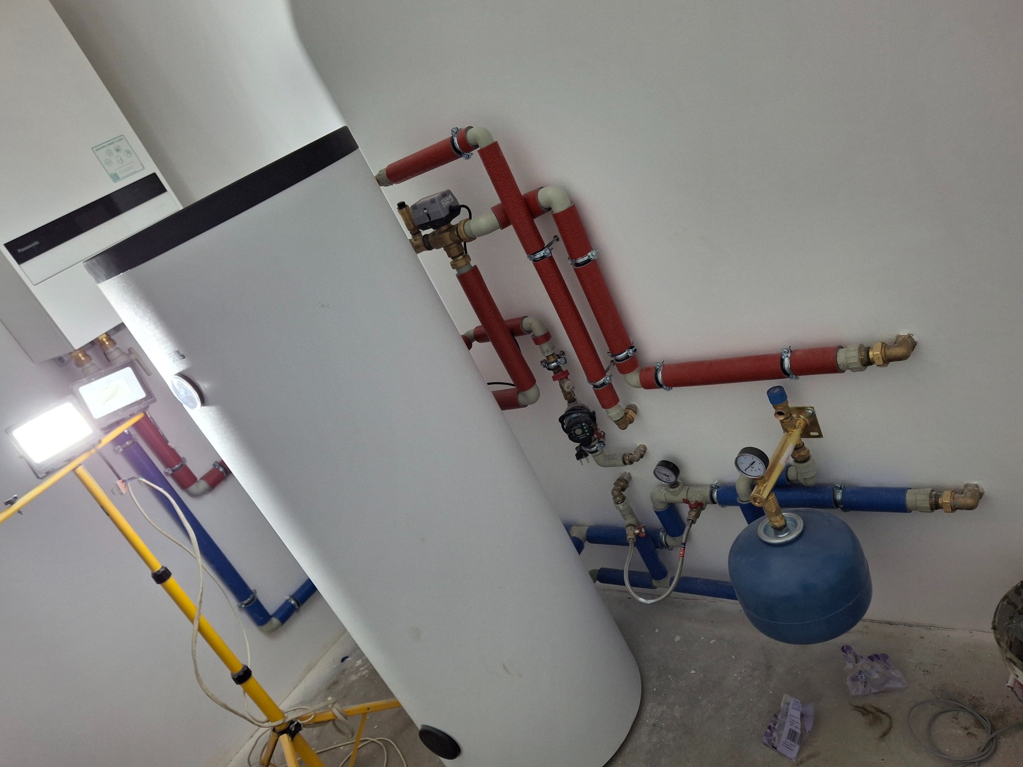Biały zasobnik ciepłej wody z podłączonymi rurami miedzianymi w izolacji i niebieskimi rurami z osprzętem, lampa budowlana oświetla instalację.