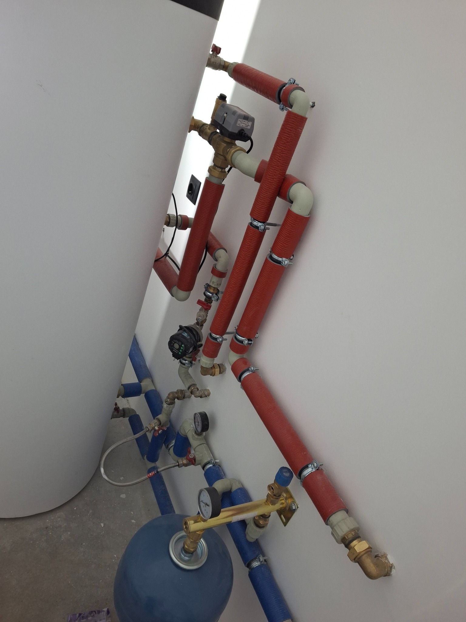 Instalacja grzewcza z czerwonymi rurami i zaworami, częściowo ukryta za białym zbiornikiem, zamontowana na białej ścianie. Widoczne manometry i elementy przyłączeniowe.